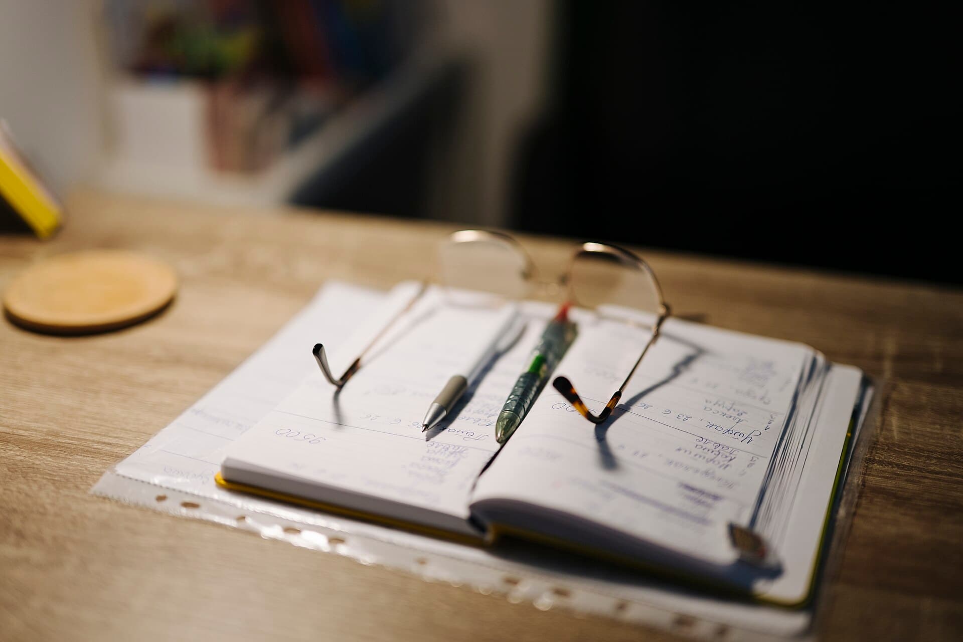 A desk with an open notebook, pens and glasses